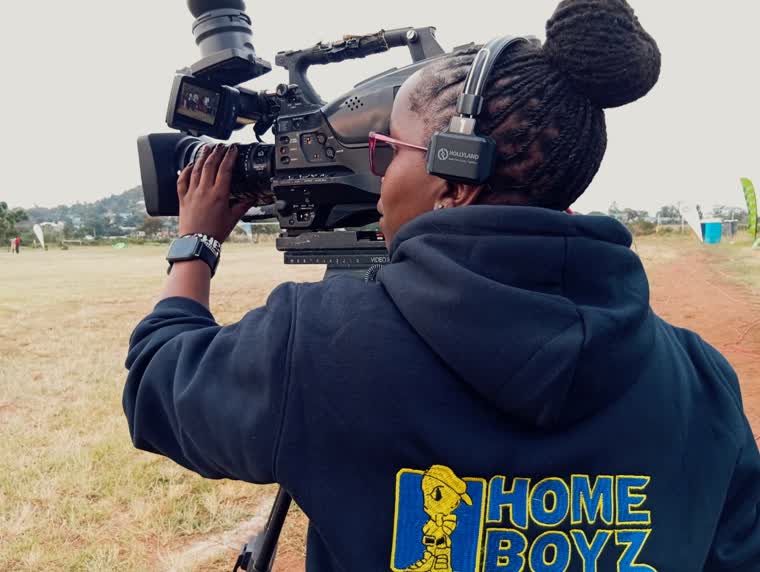 Camera operator in a Homeboyz hoodie on open ground